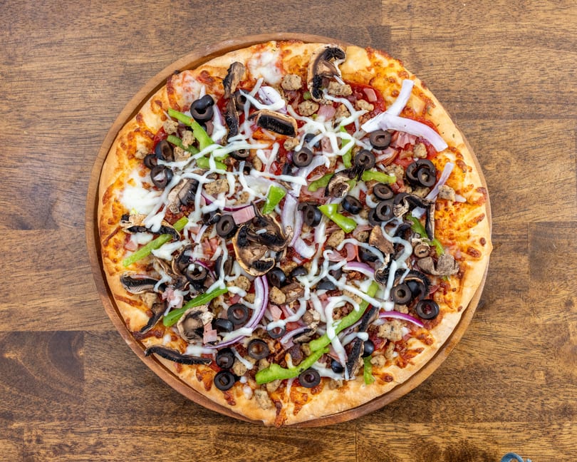 Overhead view of a supreme pizza with olives, mushrooms, peppers, onions, and sausage on a wooden board.