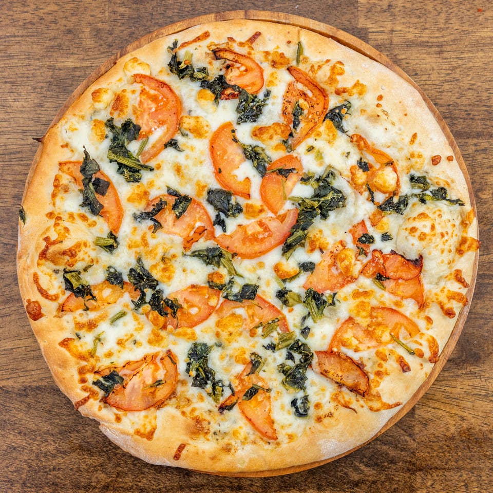 A whole white pizza with melted cheese, sliced tomatoes, and spinach on a wooden board.