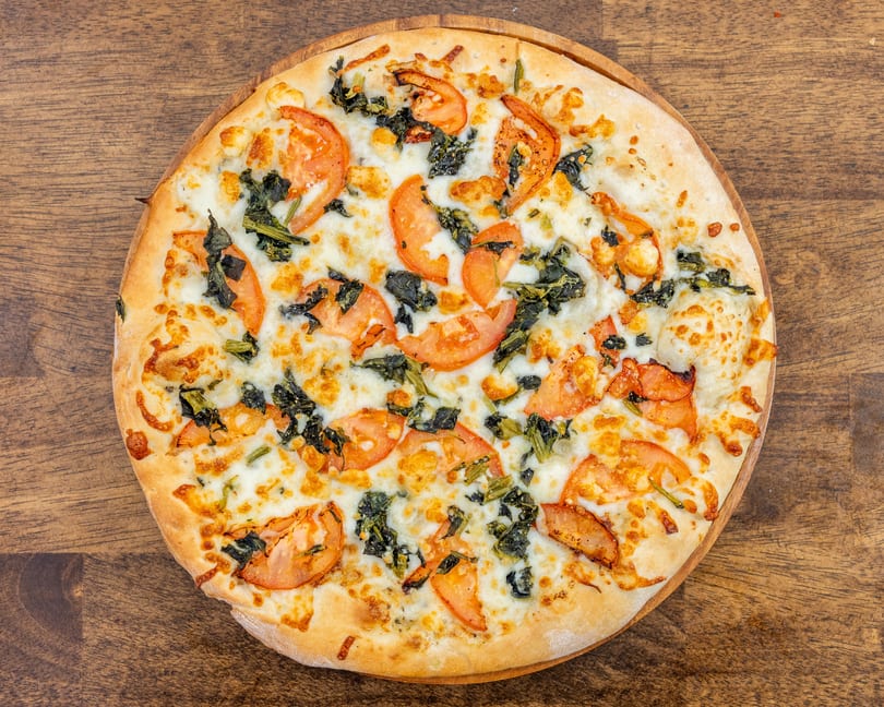 A whole white pizza with melted cheese, sliced tomatoes, and spinach on a wooden board.