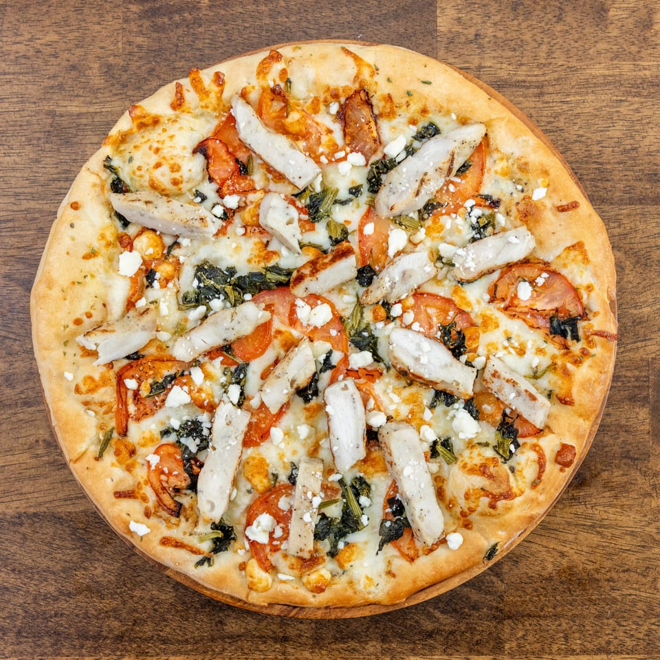 Overhead view of a chicken pizza with tomatoes, spinach, and feta cheese on a wooden table.