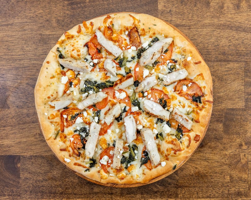 Overhead view of a chicken pizza with tomatoes, spinach, and feta cheese on a wooden table.