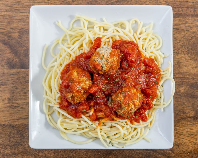 Spaghetti with meatballs and red sauce, garnished with herbs, on a white square plate.