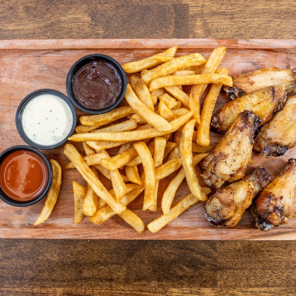 A wooden tray holds crispy french fries, roasted chicken wings, and three dipping sauces.