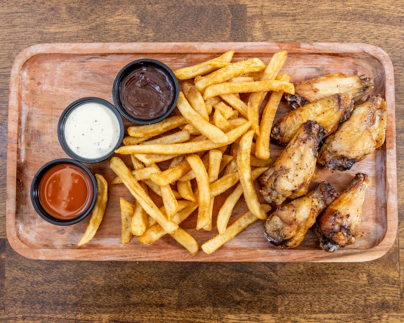 A wooden tray holds crispy french fries, roasted chicken wings, and three dipping sauces.